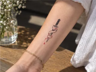 Fine line dagger with heart and ribbon forearm tattoo